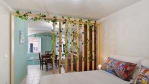 A bedroom with a bed, pillows, a wooden partition with greenery, a dining area with chairs, a kitchen in the background, and a window.