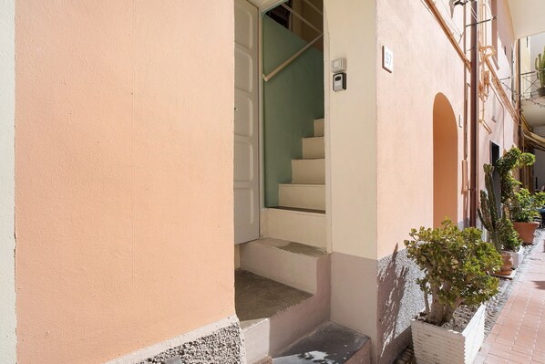 A narrow corridor with a staircase leading to an upper floor, potted plants along the path, and a door with a number sign.
