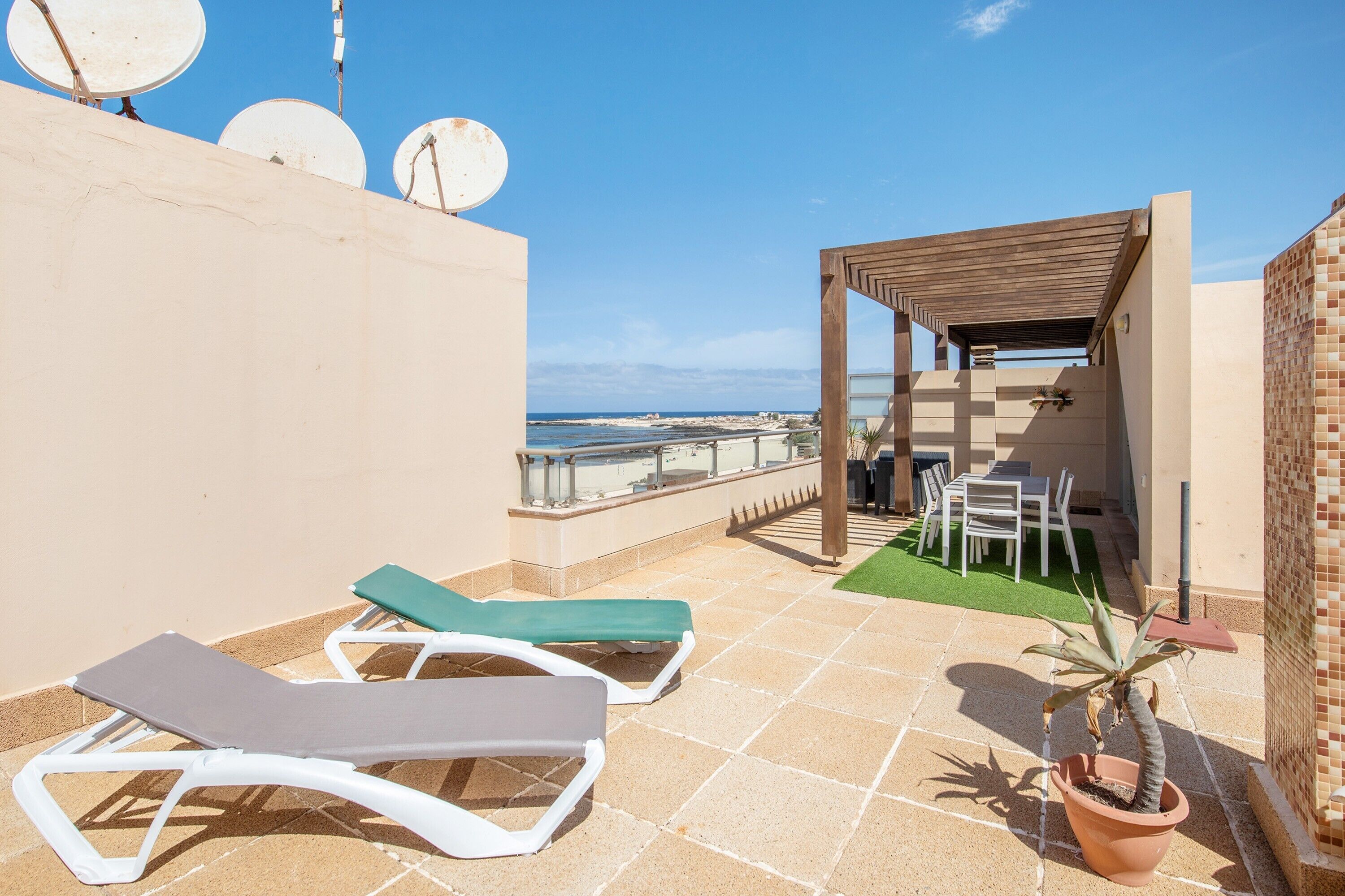 Una terraza en la azotea con sillones de descanso, una pérgola, una planta en maceta y vistas al mar.