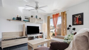 A modern living room with a flat-screen TV, wooden furniture, a ceiling fan, and a balcony with orange curtains.