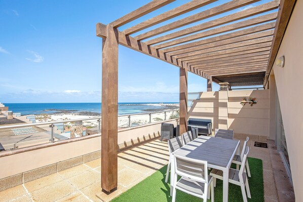 A terrace with a wooden pergola, a table with chairs, and a view of the beach and ocean.