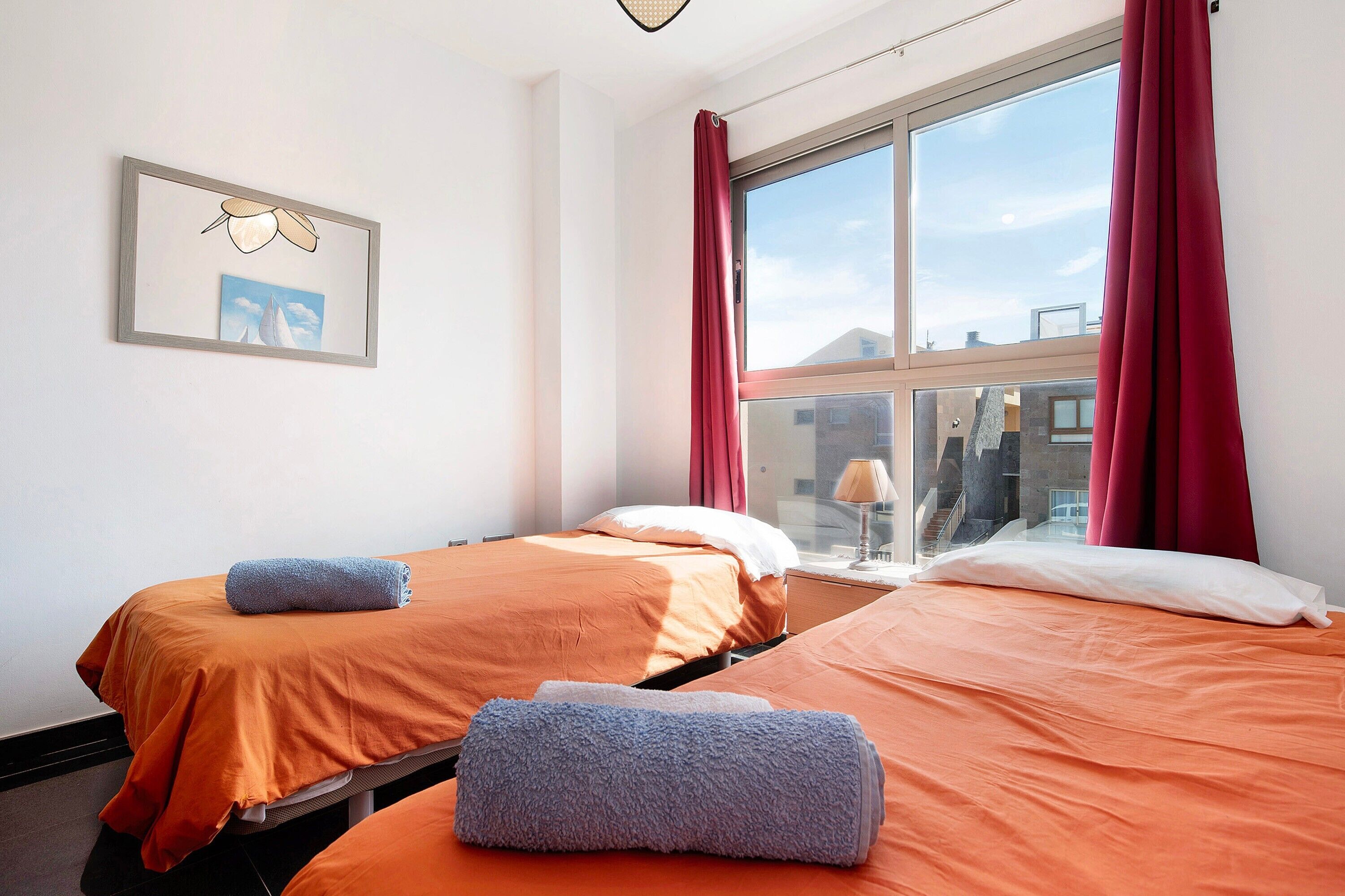 A hotel room with two beds, a window with red curtains, a framed picture on the wall, and a view of buildings outside.