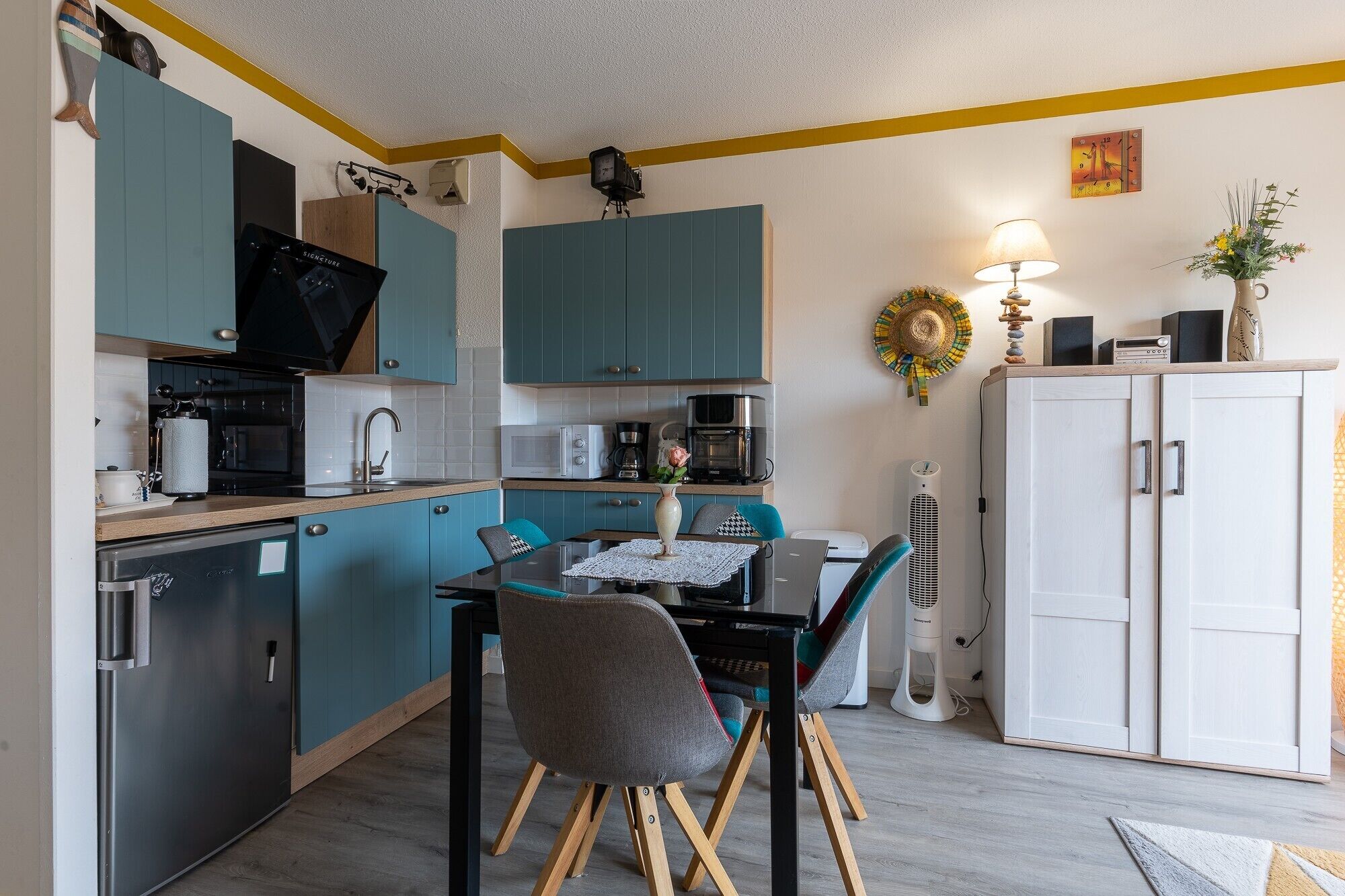A compact kitchen with teal cabinets, a dining table with chairs, a white cabinet, and a wall clock.