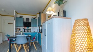 A compact kitchen with teal cabinets, a dining area with a table and chairs, and a large, textured lamp. - Studio Apartment 'Cabine Centre Ville' with Wi-Fi (ARCACHON)