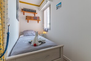Une chambre à coucher comprenant un lit, une table de chevet, une fenêtre avec des rideaux, une étagère fixée au mur avec des livres et une petite décoration murale.