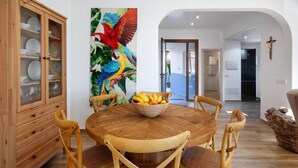 Un coin repas avec une table ronde en bois, des chaises, un panier de fruits, un buffet en bois et un tableau coloré représentant un perroquet.