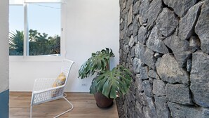 Habitación con una silla blanca, una planta en maceta, una pared de piedra y una ventana con vista a árboles.