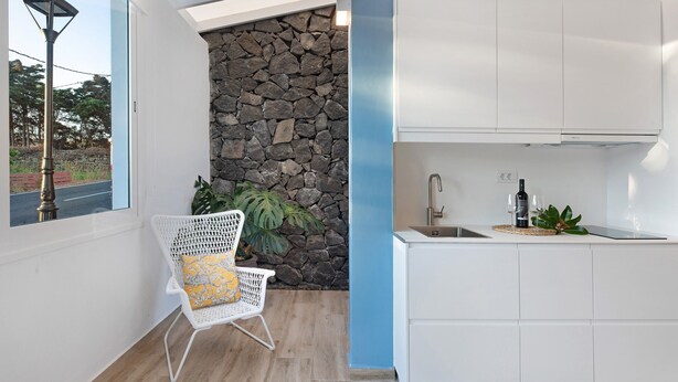 Una cocina moderna con armarios blancos, una pared decorativa de piedra, un ventanal con vistas y una silla con cojín amarillo.
