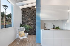 Eine moderne Küche mit weißen Schränken, einer Wand als steiniger Akzent, einem Fenster mit Ausblick und einem Stuhl mit gelbem Sitzkissen.