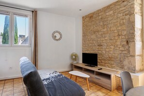 Una habitación con pared de piedra, un televisor sobre un mueble de madera, un ventilador y una ventana con cortinas.