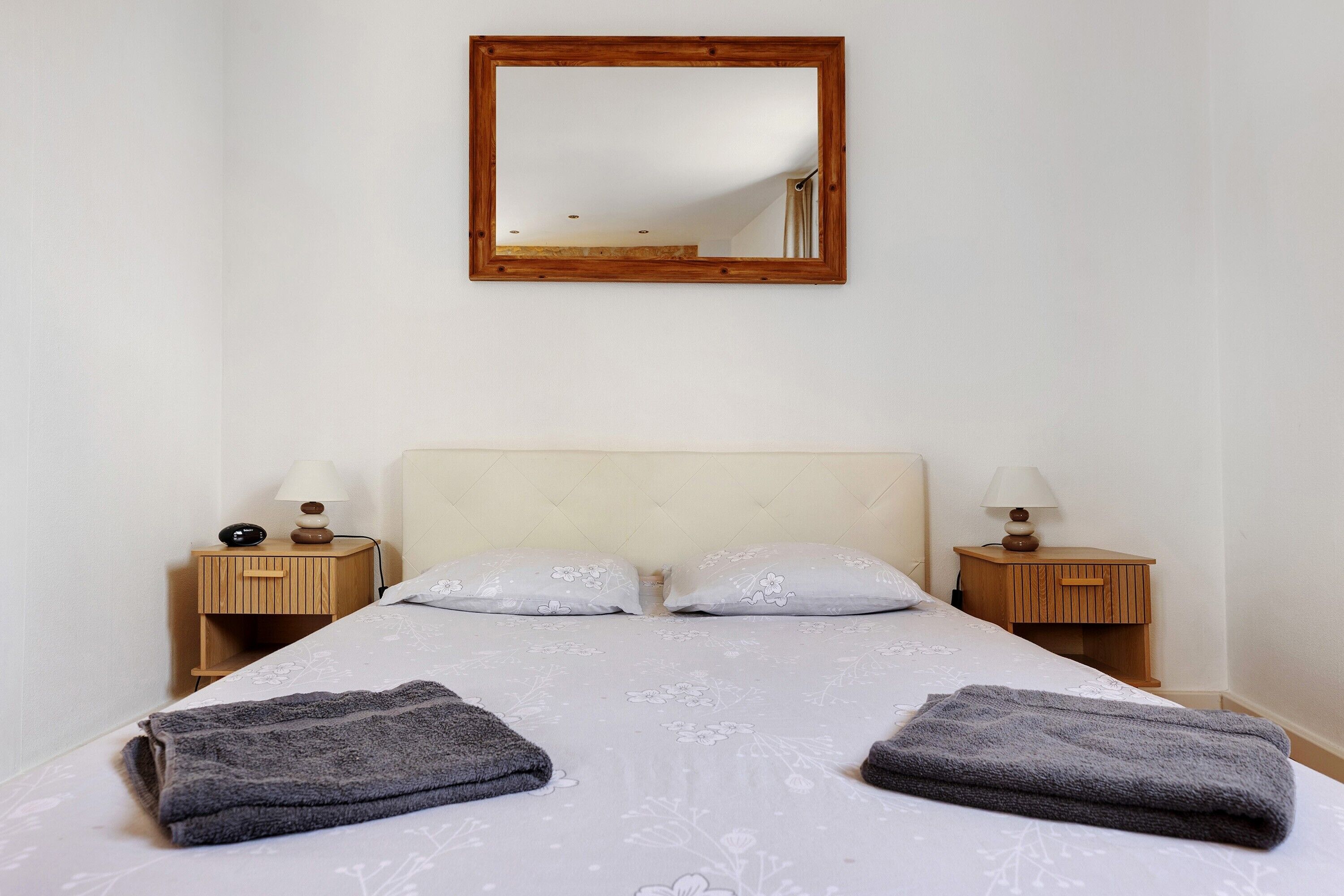 A bedroom with a bed, two bedside tables with lamps, a wooden headboard, and a mirror on the wall.