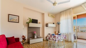 Ein Wohnzimmer mit rotem Sofa, einem Fernseher auf einem weißen Schrank, einem Esstisch mit blumigem Tischläufer und einem Balkon mit Blick.