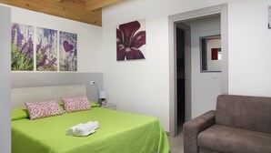 Un dormitorio con cama, colcha verde, dos almohadas estampadas, una mesita de noche con lámpara y decoración en la pared.