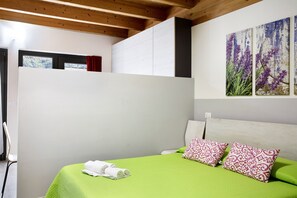 Una camera da letto con una coperta verde, due cuscini decorati, una testiera bianca e un quadro con fiori viola al muro.