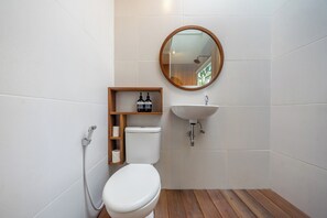 A bathroom with a white toilet, wooden floor, a wooden shelf with toiletries, a round mirror, and a white sink. - ZenDen (Tibubeneng)