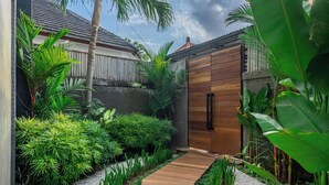 A garden pathway leading to a wooden door, surrounded by lush greenery and a concrete wall. - ZenDen (Tibubeneng)