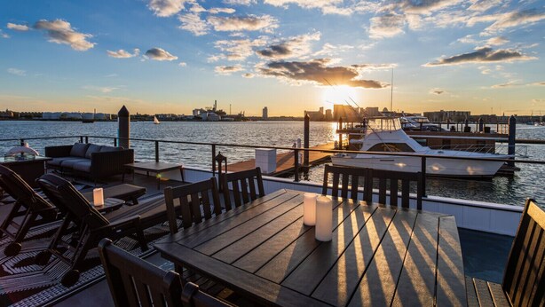 A waterfront patio with outdoor seating, a view of the sunset over the water, and a marina with boats.