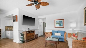 A living room with a blue sofa, a wooden coffee table, a TV mounted on the wall, and a ceiling fan. - Ocean Views W/Full-Kitchen Await - Kaha Lani #104 (LIHUE, KAUAI)