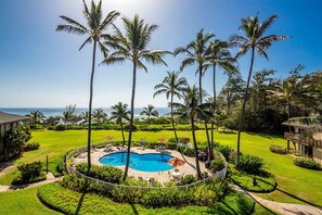 A circular pool surrounded by palm trees, a lawn, and a building with a balcony. - Ocean Views W/Full-Kitchen Await - Kaha Lani #104 (LIHUE, KAUAI)