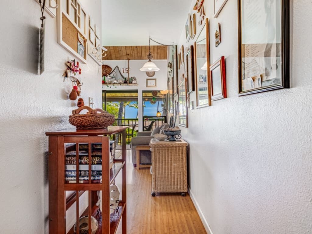 Un couloir étroit où se trouve une table en bois sur laquelle sont exposés des livres, ainsi qu’un panier en osier. Les murs sont ornés de tableaux encadrés et d’un grand miroir.