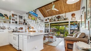 Una cocina amplia con armarios blancos, una isla central y un comedor con sofá marrón. El techo es de madera con un ventilador, y la cocina tiene un ventanal que ofrece vistas al exterior.