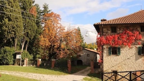 Un bâtiment rustique en pierre, doté d’un toit en tuiles, un chemin de terre menant à une petite construction et un arbre aux feuilles d’automne.