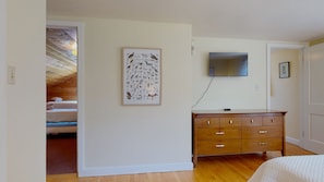 A bedroom with a wooden dresser, a framed bird illustration, a flat-screen TV, and a staircase leading to an attic.