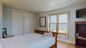 Un dormitorio con cama, una silla, armario, ventana con vista al mar y televisión en la pared.