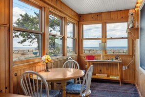 Una habitación con paneles de madera, una mesa y sillas, ventanas amplias con vista al mar y una lámpara sobre la mesa.