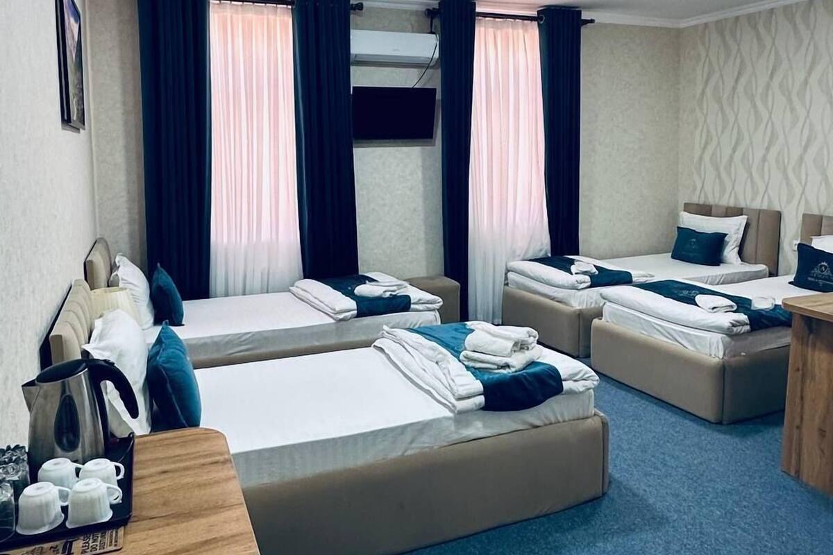 Habitación de hotel con dos camas individuales, un sofá cama, una mesa de madera con tazas y una tetera, y un televisor de pantalla plana montado en la pared.
