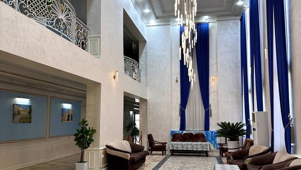 A spacious lobby with a high ceiling, a chandelier, blue curtains, and a seating area with sofas and a coffee table. - AFSONA HOTEL (Bukhara)