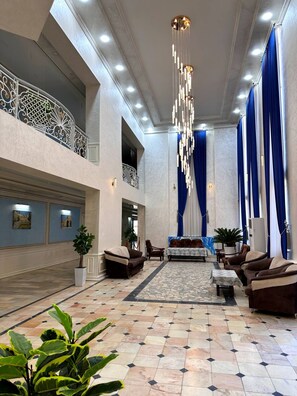 A spacious lobby with a high ceiling, a chandelier, blue curtains, and a seating area with sofas and a coffee table. - AFSONA HOTEL (Bukhara)