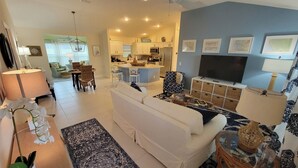 A modern living room with a white sofa, a dining area with a table and chairs, a kitchen with white cabinets, and a TV on the wall. - After Dune Delight - 3/2 w/ Golf Cart in The Village of Cason Hammock (Cason Hammock, The Villages)