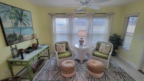 A living room with a green sofa set, a small round table with a lamp, a painting of palm trees, and a patterned rug. - After Dune Delight - 3/2 w/ Golf Cart in The Village of Cason Hammock (Cason Hammock, The Villages)