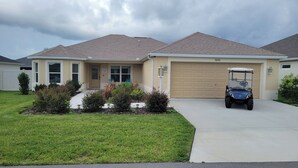A single-story house with a beige garage door, a driveway, and a golf cart parked in front. - After Dune Delight - 3/2 w/ Golf Cart in The Village of Cason Hammock (Cason Hammock, The Villages)