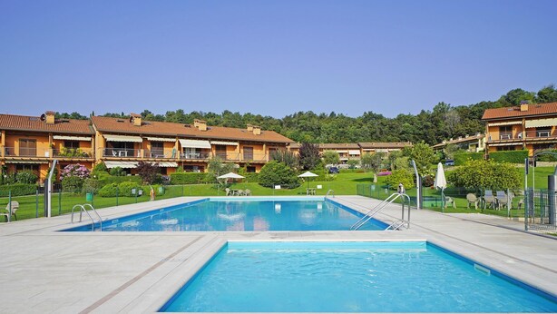 Ein Schwimmbecken mit Fliesenplattform, umgeben von einem Zaun. Im Hintergrund sind Gebäude mit Balkonen und eine hügelige Landschaft zu sehen.
