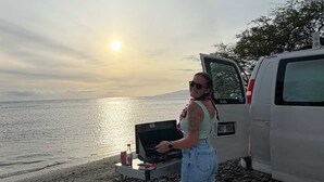 A person at a beach with a laptop on a portable table, a surfboard, and a van nearby. - Deluxe Camper Van  with Aloha (Kihei)