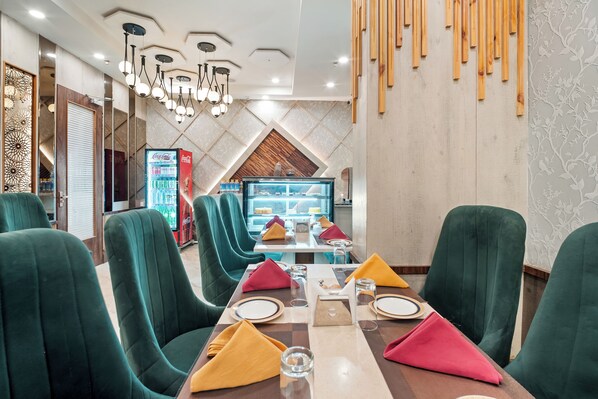 A restaurant with green upholstered chairs, a central dining table set with plates, glasses, and napkins. A Coca-Cola refrigerator is visible in the background.