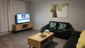 A living room with a wooden coffee table, a black leather sofa, a TV on a wooden stand, and a painting on the wall.