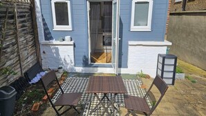 Una casa azul con puerta y ventanas blancas, un patio con piso a cuadros y dos sillas metálicas con una mesa.