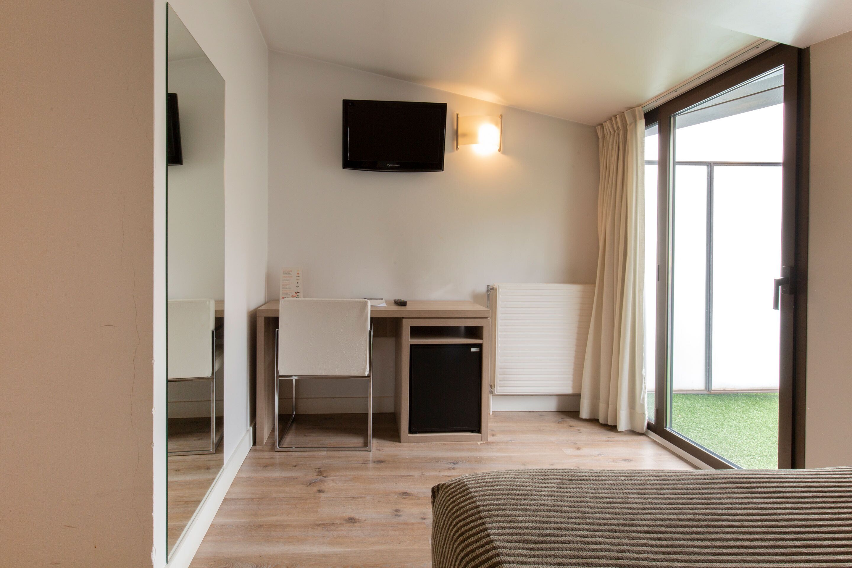 A hotel room with a bed, a desk with a chair, a television mounted on the wall, and a sliding glass door to the outside.