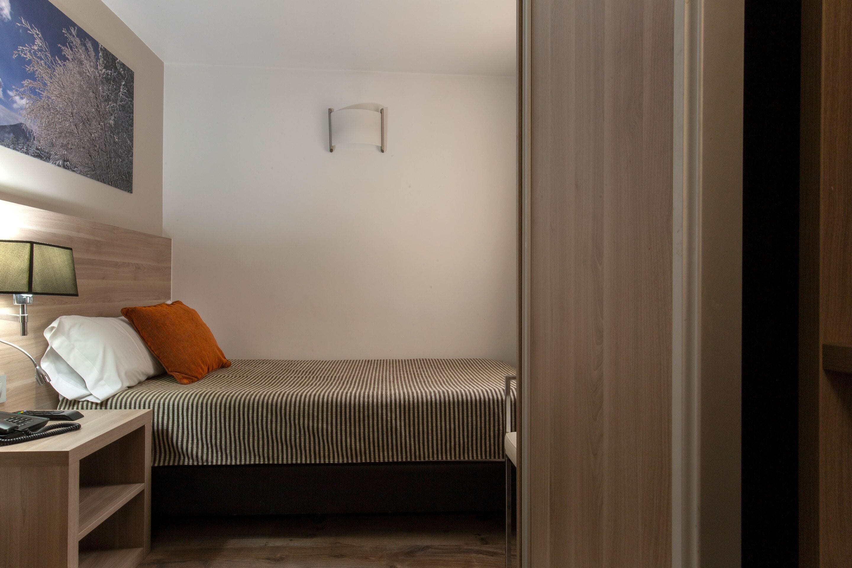 A compact hotel room with a single bed, a bedside table with a lamp, a small shelf, and a wall-mounted light fixture.