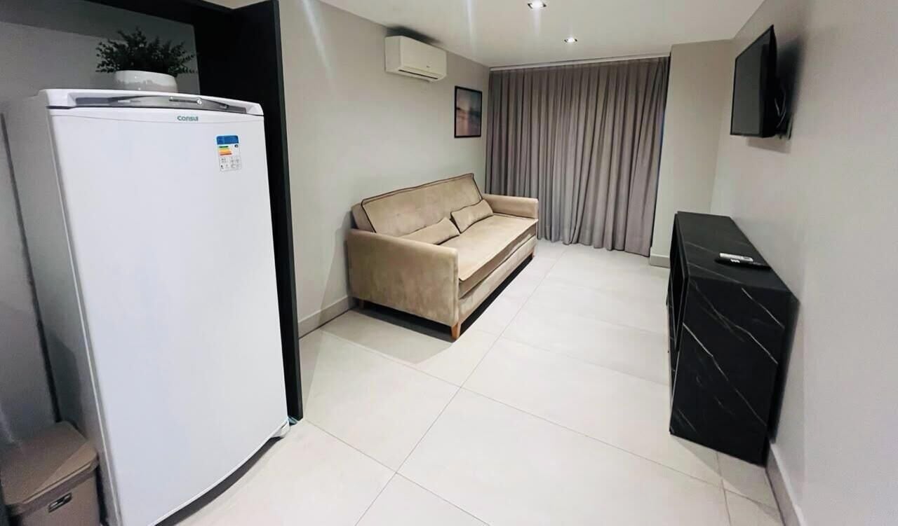 Un salon avec un canapé beige, un meuble de rangement noir pour appareils électroniques et un réfrigérateur blanc.