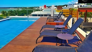 Un espace piscine aménagé sur le toit, avec des chaises longues et des tables, offrant une vue dégagée sur la mer et des bâtiments en arrière-plan.