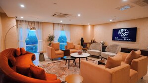 A modern living room with a curved orange sofa, beige armchairs, and a coffee table. A flat-screen TV is mounted on the wall, and there is a large window with sheer curtains. - Nine Tree Luxury Hotel & Suites (Lahore)