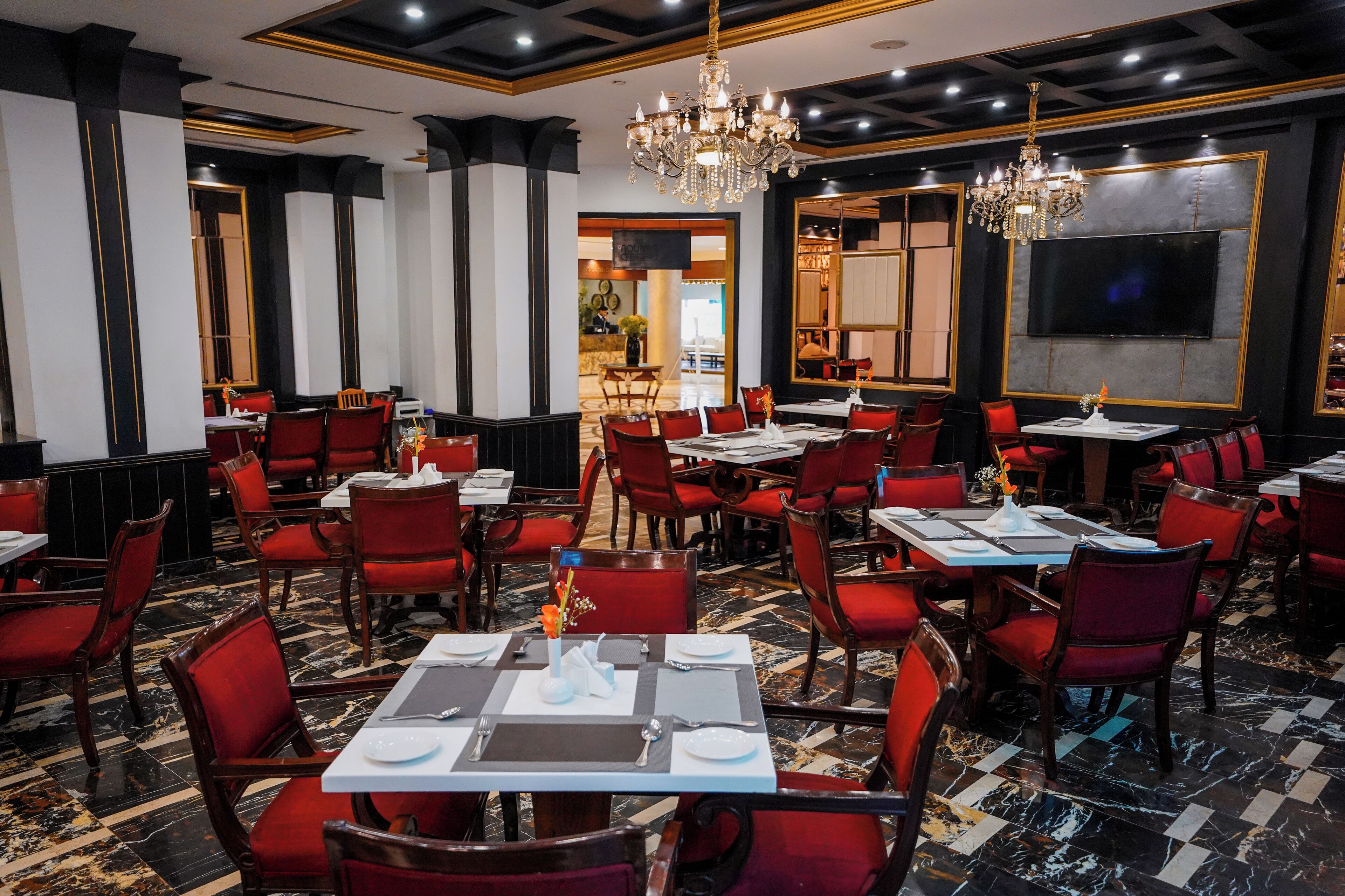 Un restaurant avec des chaises à revêtement rouge, un sol en marbre, des lustres et un téléviseur à écran plat fixé au mur.