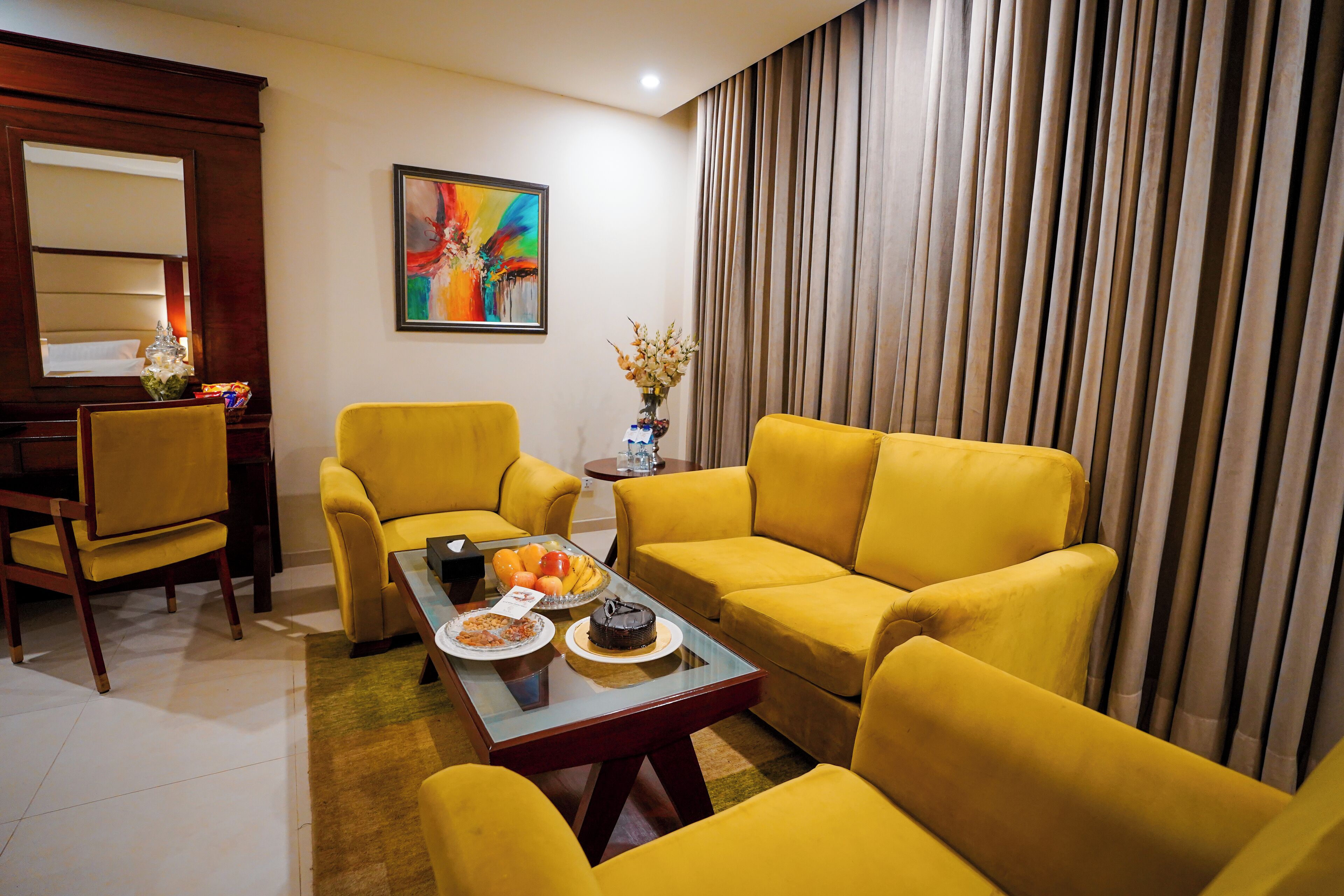 A modern living room with yellow sofas, a glass coffee table, and a dining table with a cake and fruit.