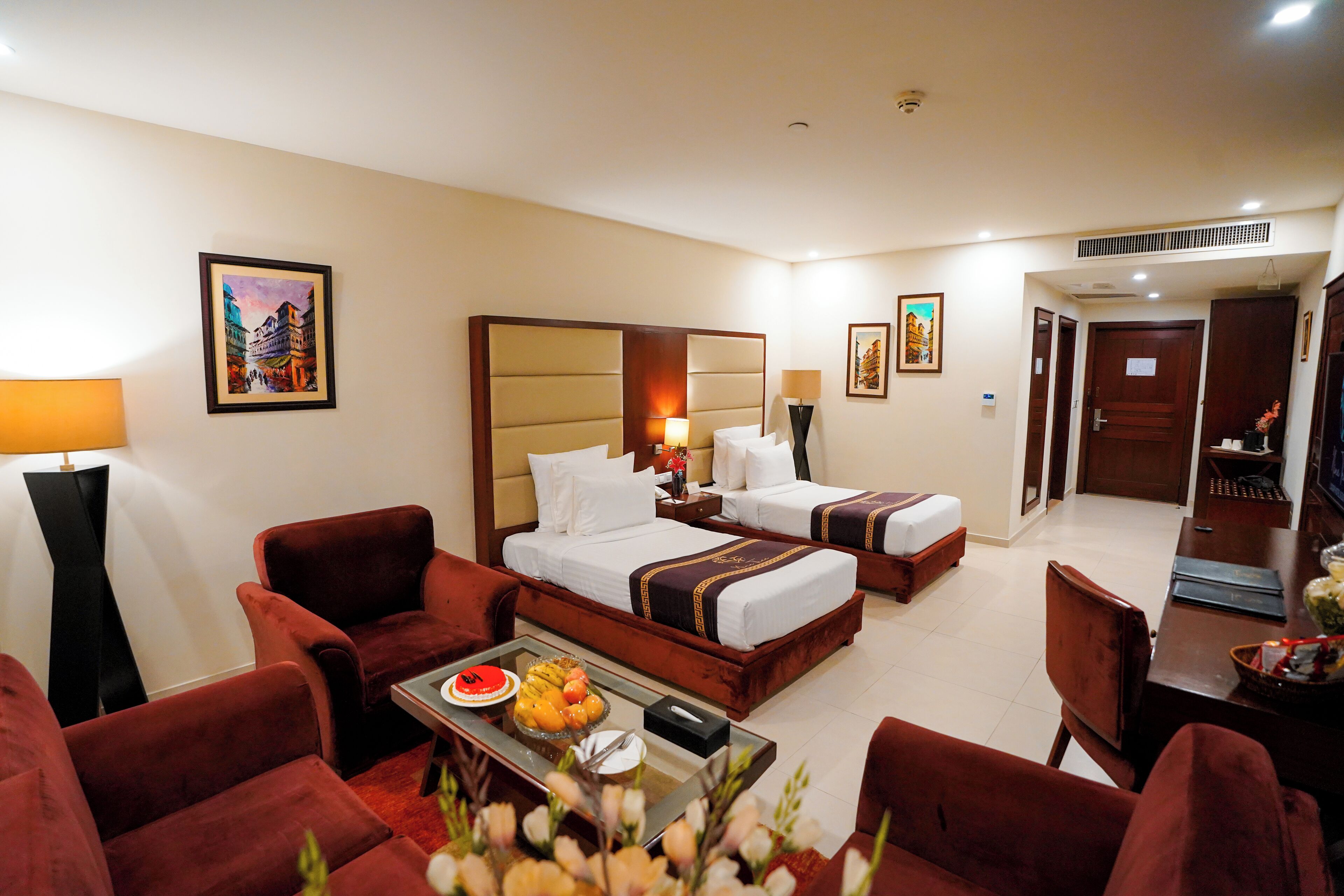 A hotel room with two beds, a sitting area with red chairs, a dining table with fruit, and artwork on the walls.
