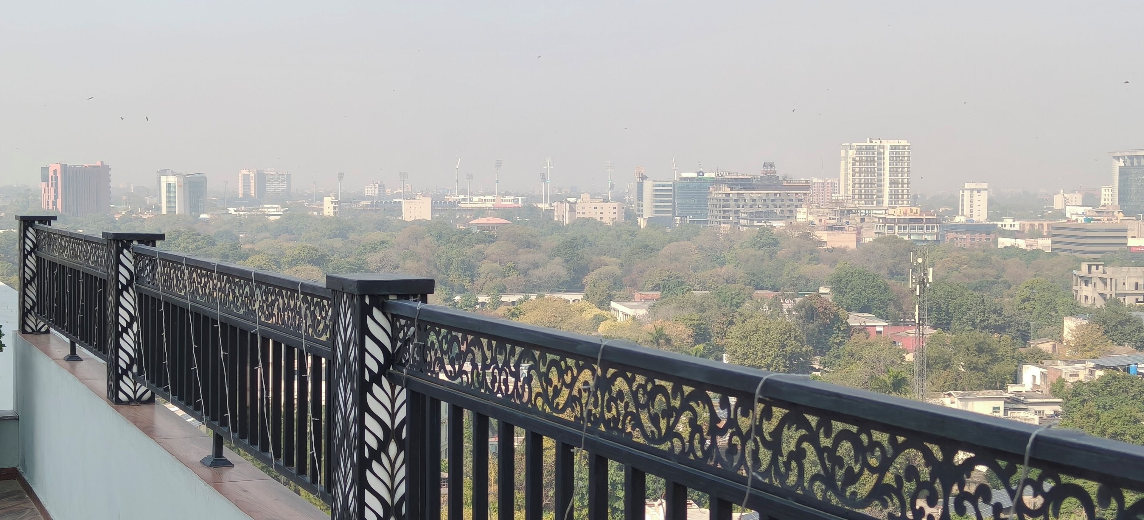 A balcony with ornate black railings overlooking a cityscape with various buildings and greenery.