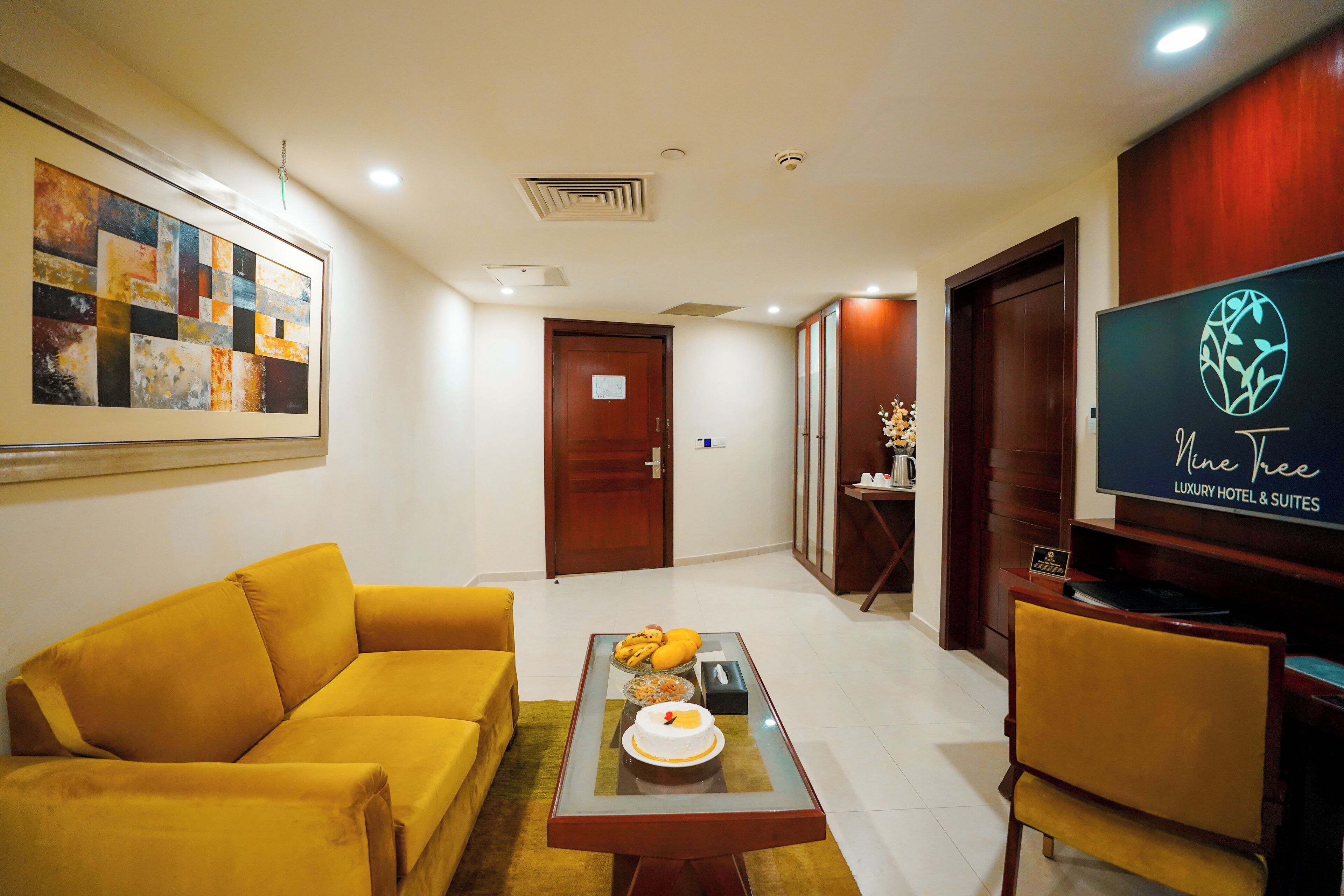 A modern living room with a yellow sofa, a glass coffee table with fruits and a cake, a flat-screen TV, and a framed abstract painting.